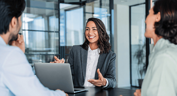 A businesswoman smiling while speaking with colleagues around a laptop in an office meeting.