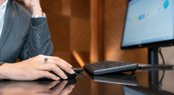 Close-up of a hand using a computer mouse at a desk with a monitor showing charts.